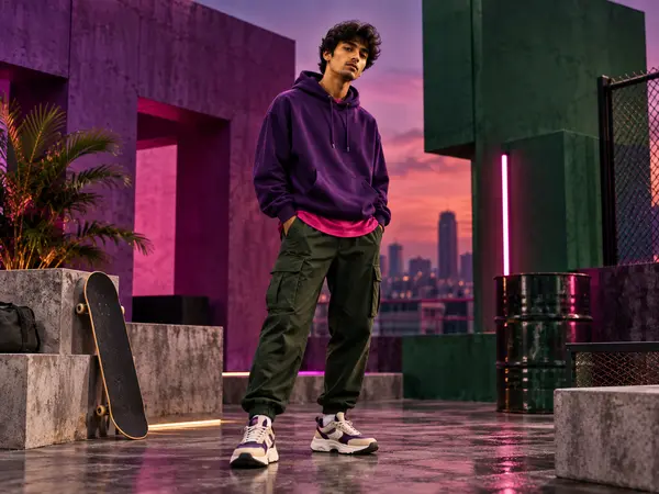 Young man in a purple hoodie and green cargo trousers with a skateboard on a rooftop at dusk
